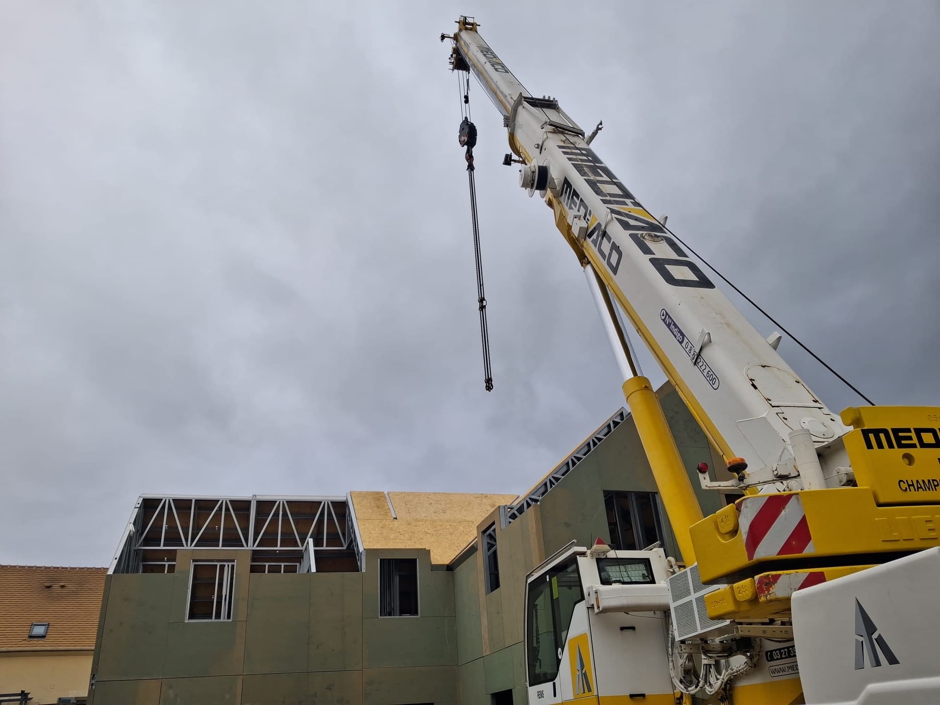Levage à la grue d'éléments d'ossature métallique légère : fermes de toiture en acier et plancher, sur un bâtiment en construction accolé à l'existant